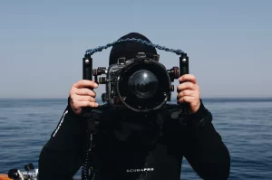 Photograph of a man in a scuba diving suit holding an underwater camera hanging on the edge of a boat