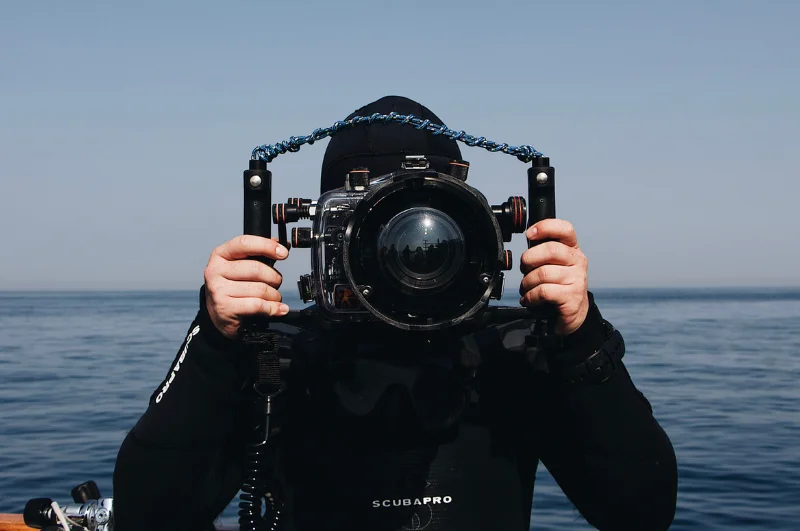 Photograph of a man in a scuba diving suit holding an underwater camera hanging on the edge of a boat