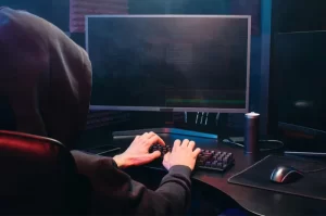 Photo of a hooded hacker or scammer sitting at a desktop computer