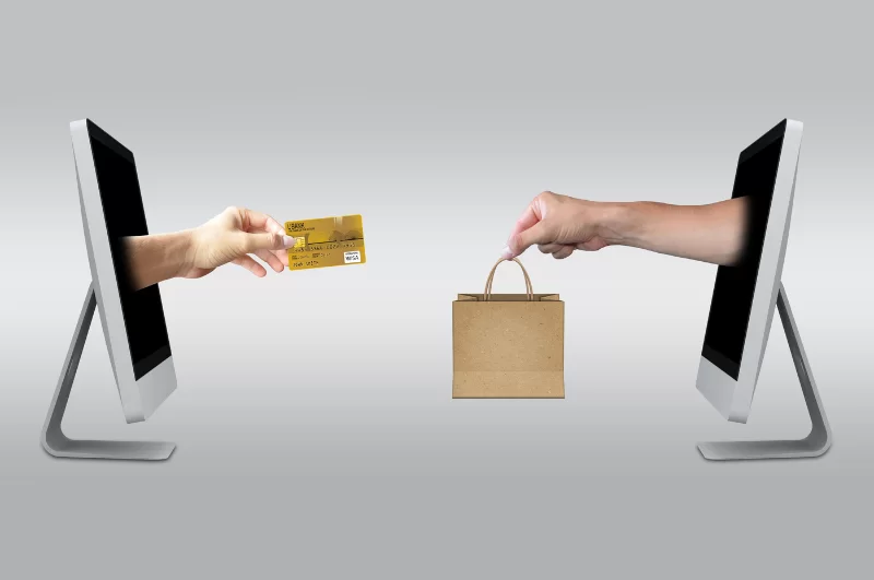 Image of hands coming out of computer monitors with one holding credit card and another holding bag representing online shopping