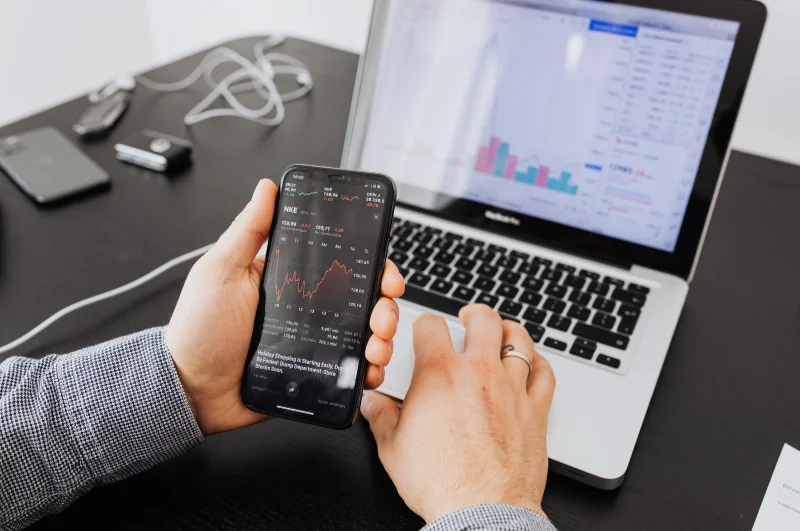 Photograph of a man holding a cell phone with a laptop tracking stock prices