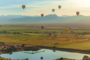 Audrey Cramer Photography's fine art aerial photograph of three hot air balloons flying over a field and reflecting in a pond in Panguitch Utah with a blue partly cloudy sky