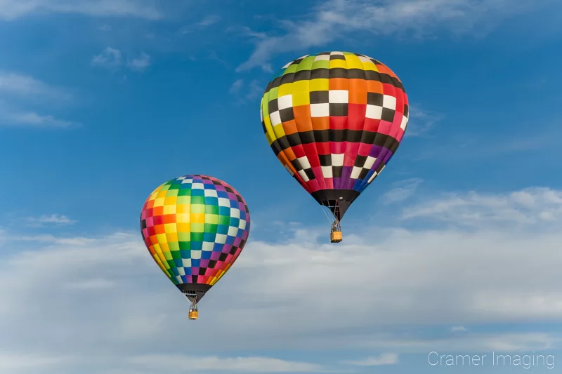 Audrey Cramer Photography's fine art photograph of two rainbow-colored hot air balloons taking flight in Panguitch Utah with a blue partly cloudy sky