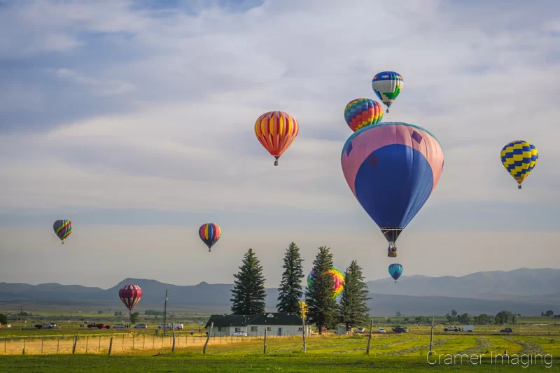 Audrey Cramer Photography's fine art photograph of a hot air balloon cluster drifting over a field in Panguitch Utah with a blue partly cloudy sky