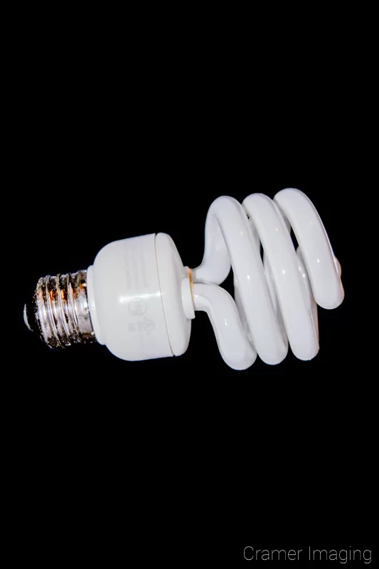 Photograph of a single compact fluorescent light bulb on its side on a black background by Audrey Cramer Photography