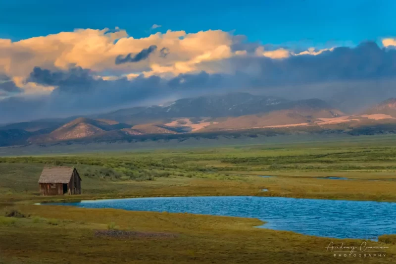 Audrey Cramer Photography's fine art landscape photograph of an abandoned lonely cabin by a rippled pond with mountains and dramatic clouds