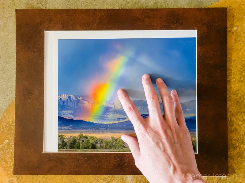 Photo of a hand adjusting the position of Audrey Cramer Photography's landscape photo titled "Broken Rainbow" inside the window of a hinged brown mat