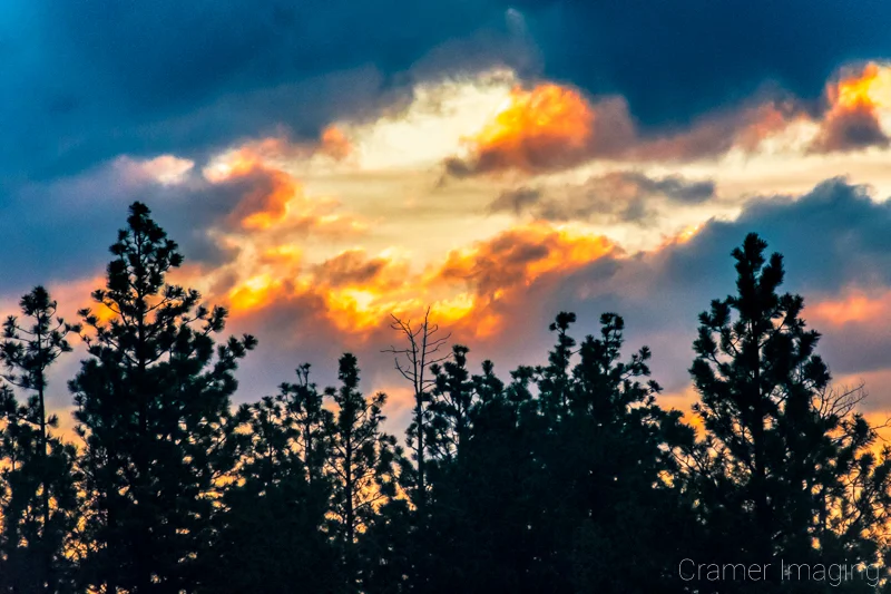 Cramer Imaging's fine art landscape photograph of a fiery golden sunset peaking out from a hole in clouds with Utah trees in silhouette