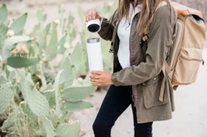 Photograph of a woman wearing backpack, standing next to cactus, opening water bottle