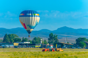 Audrey Cramer Photography's fine art photo of a hot air balloon hovering over a farm field with tractor and a town in the background