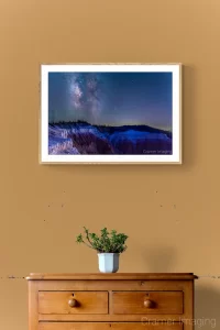 Photo of Audrey Cramer Photography's "Cedar Breaks Milky Way" on a brown wall with holes and paint scuffs with a wooden chest of drawers and a potted plant