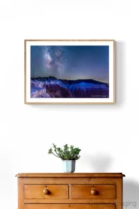 Photo of Audrey Cramer Photography's "Cedar Breaks Milky Way" on a white wall with a wooden chest of drawers and a potted plant